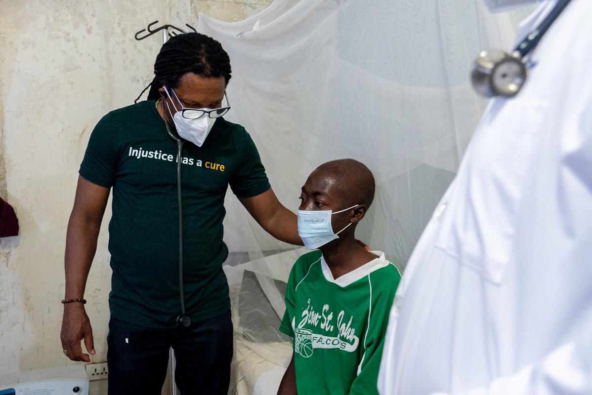 Dr. Maxo Luma, in a face mask and t-shirt that says "injustice has a cure," stands with a patient in a green shirt, also wearing a face mask.