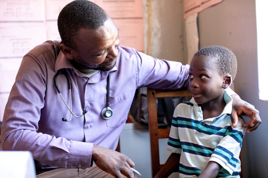 At PIH-supported Lisungwi Community Hospital in Neno, Malawi, Clinical Officer Medson Boti sits with 7-year-old Kevini Jamu, who receives free, specialized care and medicines for sickle cell disease.