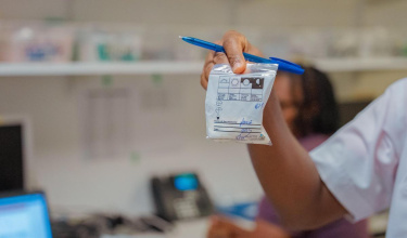a hand with a blue pen holds up a small white plastic bag containing medication