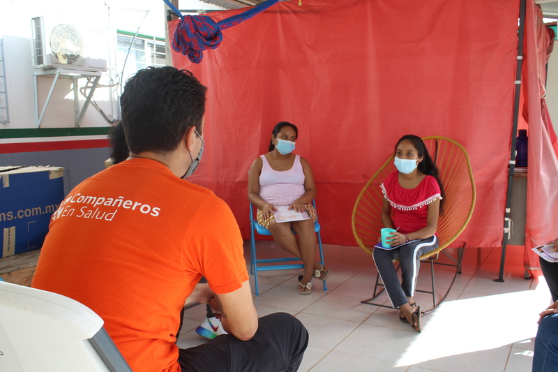 A PIH worker in an orange shirt that reads Compañeros En Salud meets with two women in Reforma, a community in Chiapas, Mexico where PIH works.