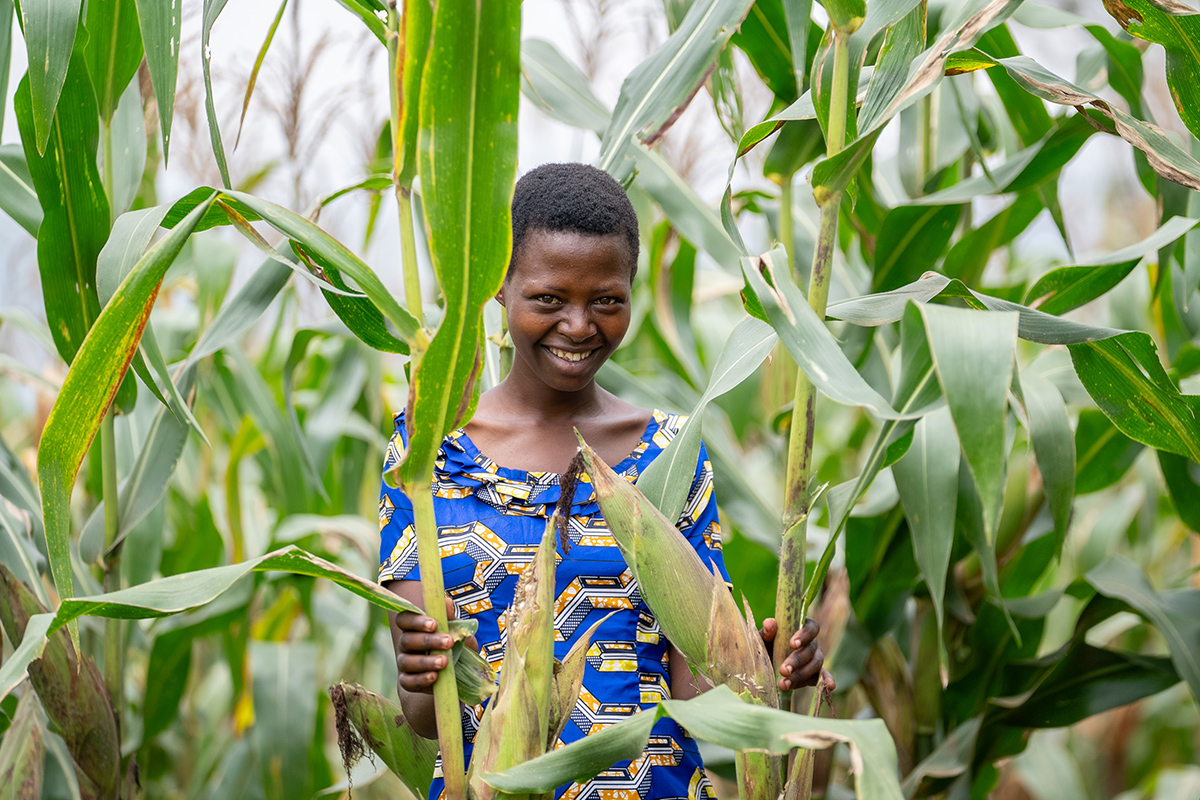 Uwizeyimana Garas stands amidst tall maize, smiling in a blue dress