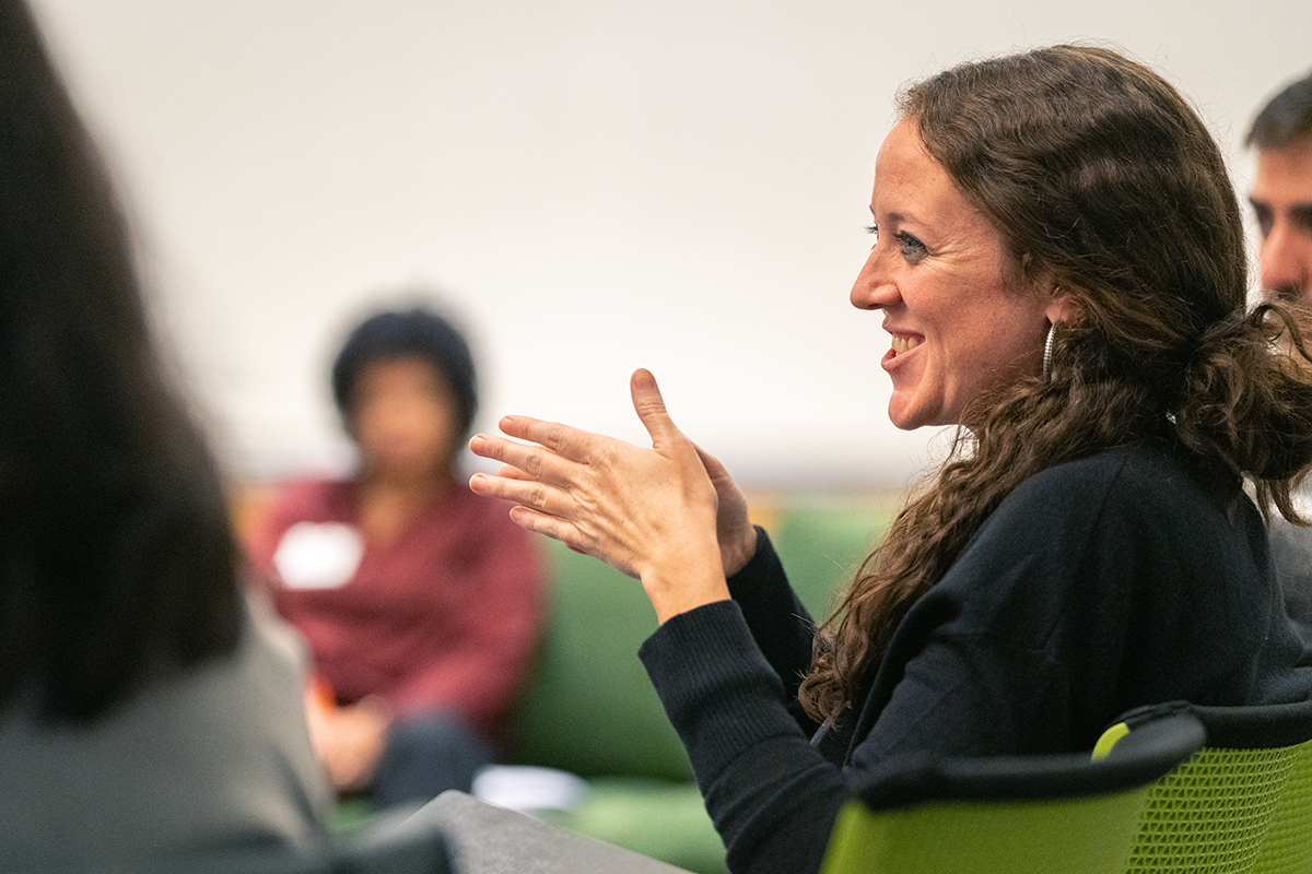 Katie Bollbach, sitting in a green chair in the right side of the frame, smiles and moves her hands as she speaks with others.  