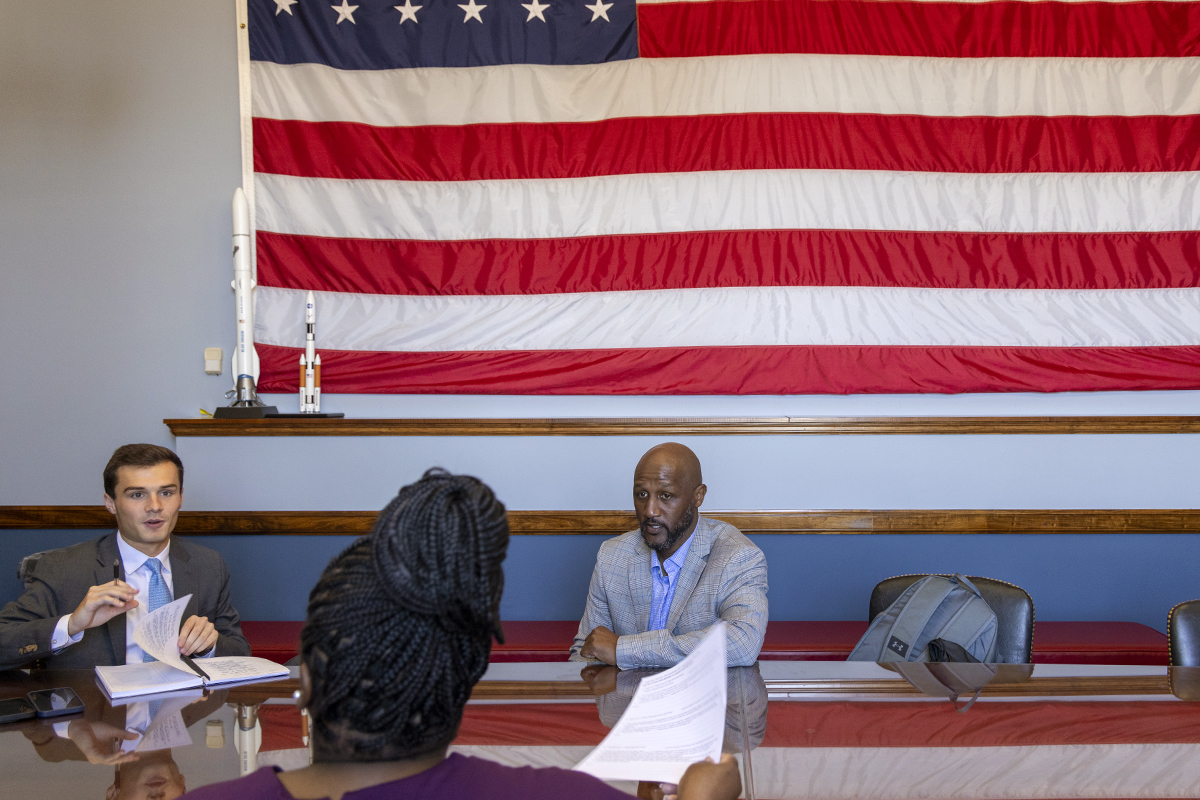 Three people talk with one another while sitting at tables underneath an American flag.