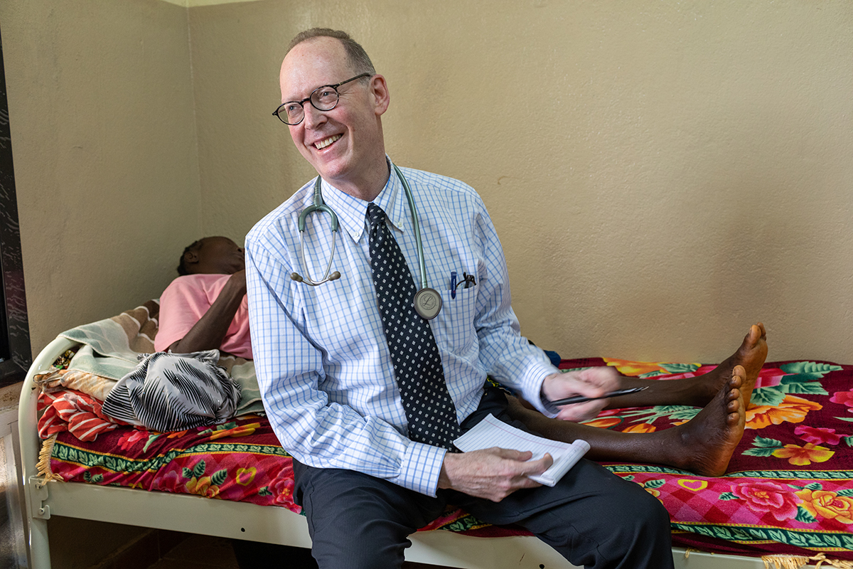 A man in glasses wearing a blue shirt, dark tie, and dark pants with a stethoscope around his neck sits on the edge of a hospital bed with colorful sheets and smiles