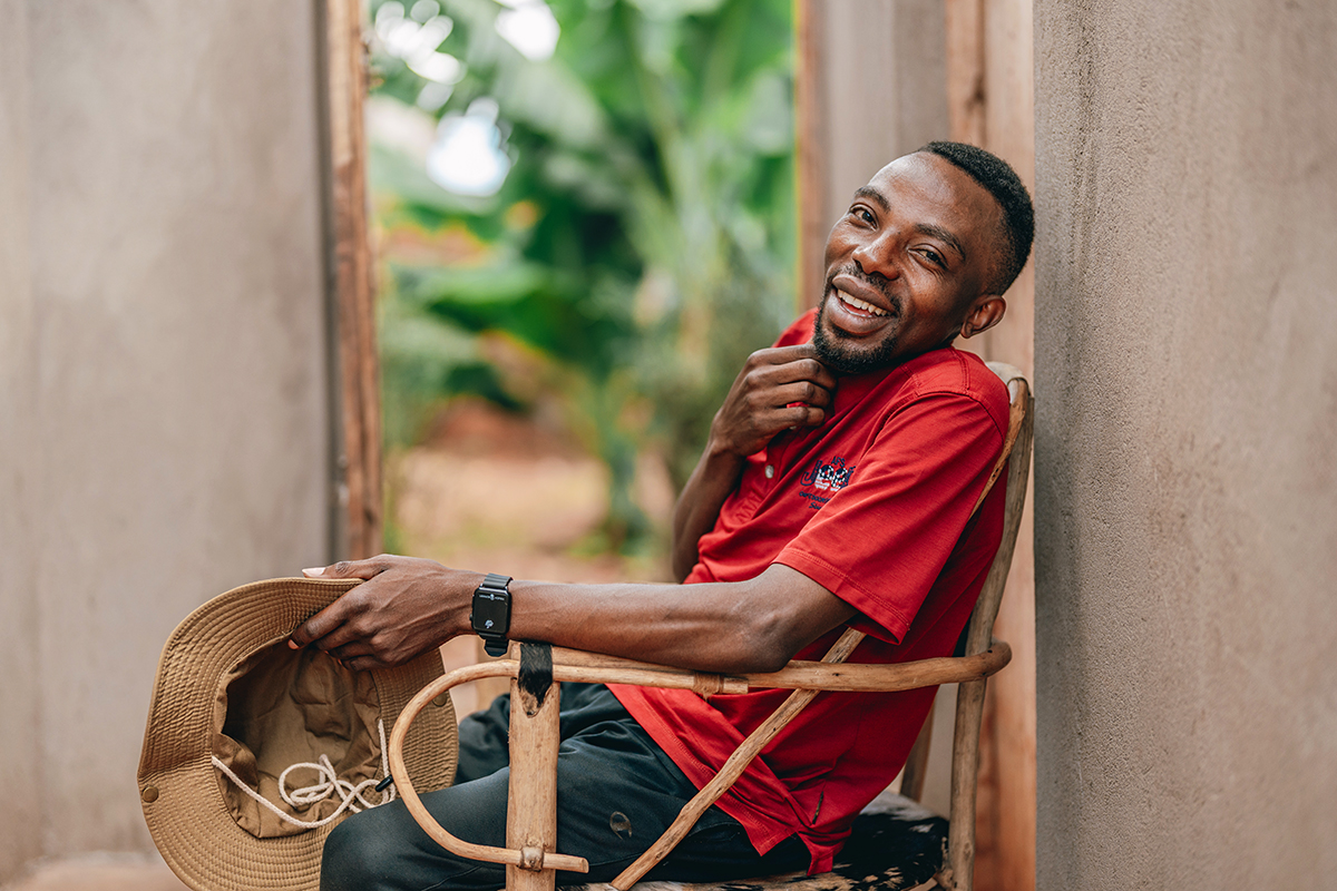 A man in a bright red shirt sits in a wooden chair, smiling. His right hand is brushing his chin, and his left hand is clutching a hat.