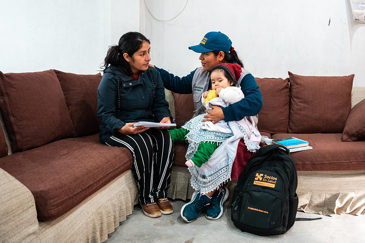 a person with a blue Socios En Salud hat holds a child wrapped in blankets and puts her hand on the shoulder of a woman to her right holding papers