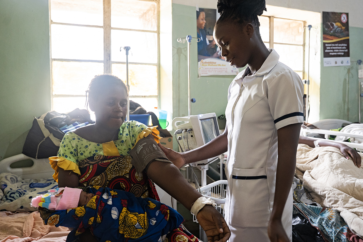 a nurse stands beside a partient sitting with a baby in her lap with a blood pressure cuff around her upper arm