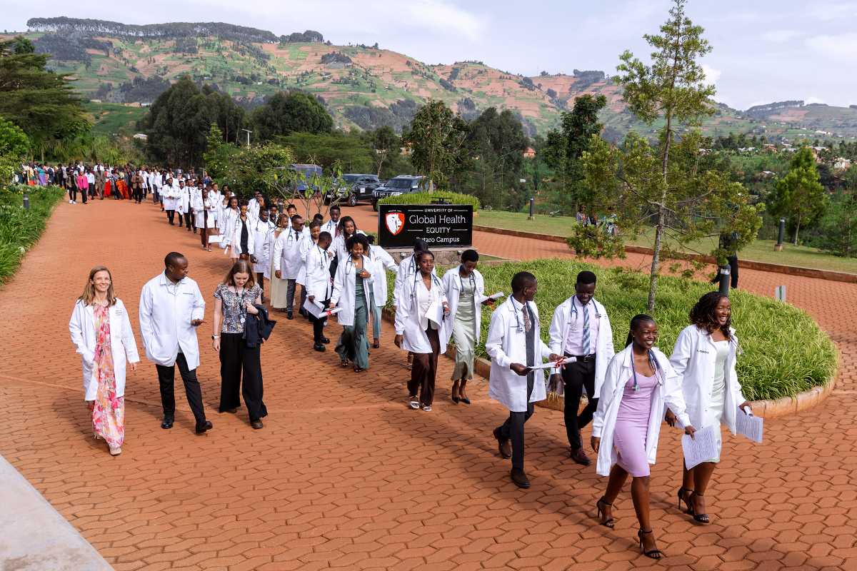 A long line of students in white coats and formal attire walk along a road.