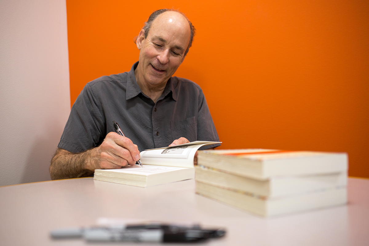 Tracy Kidder sits at a round table against an orange wall signing books with a sharpie with a smile on his face