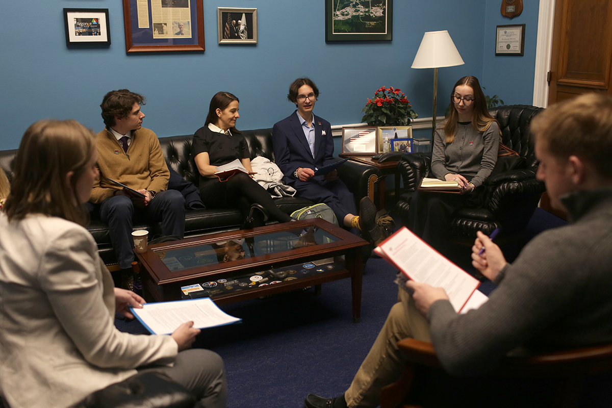 In a room with blue walls, several people sit on a leather couch and chairs for a meeting