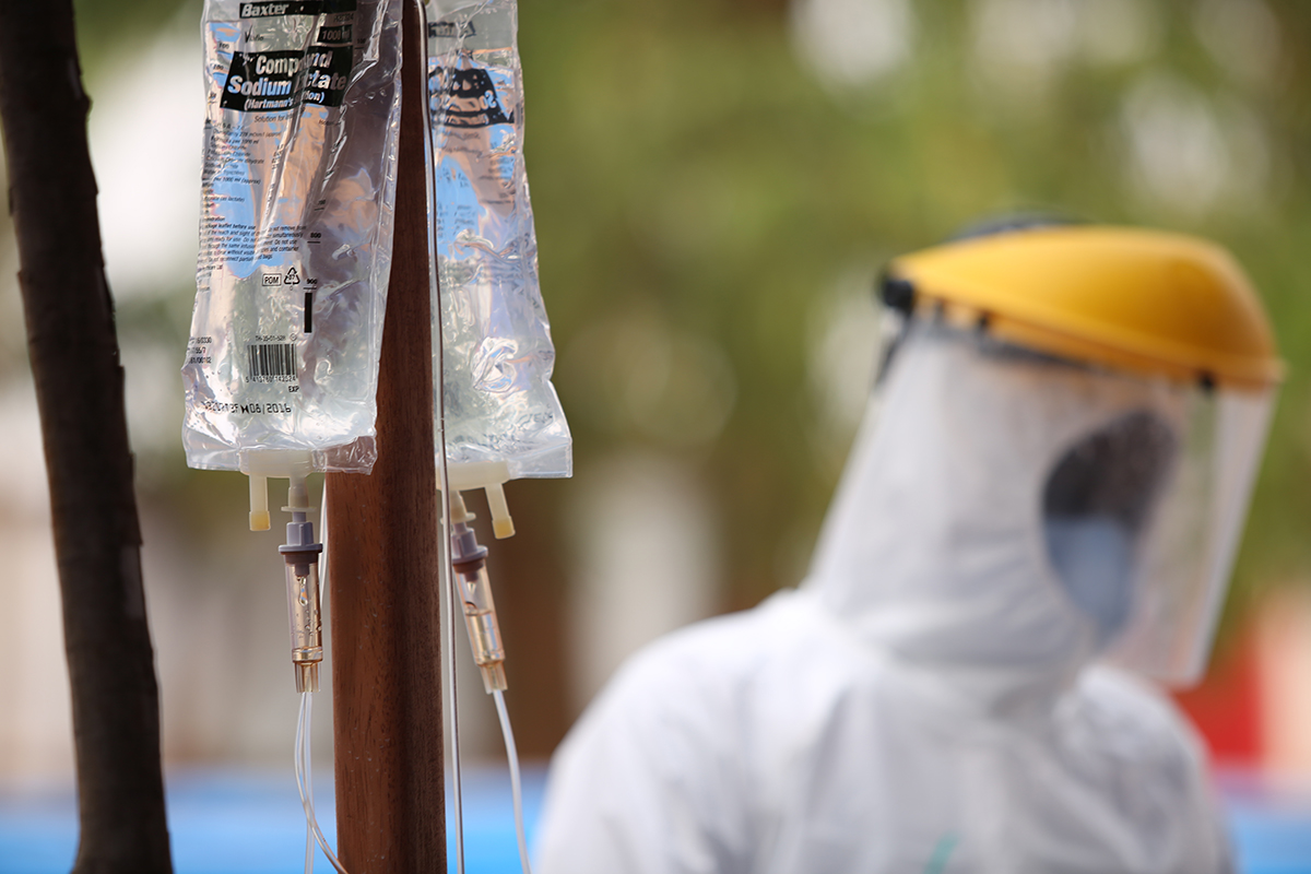 Image shows a clear medical bag hooked up to drip. In the background, there is a blurred health care worker wearing white protective equipment and a mask.