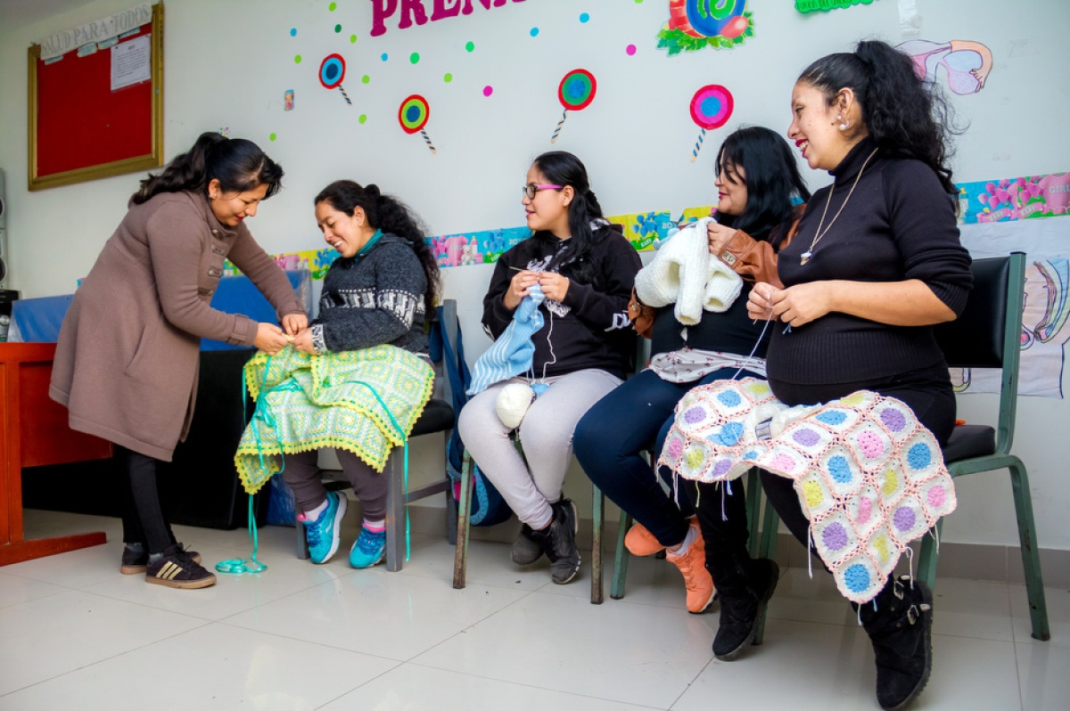 In Peru, mothers sit in a semicircle with knitted blankets and clothes on their laps as an instructor helps them.