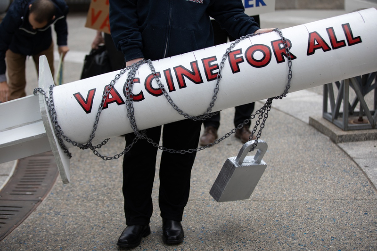 an oversized, white prop vaccine with gray chains and a lock around it that say "VACCINE FOR ALL" in red 
