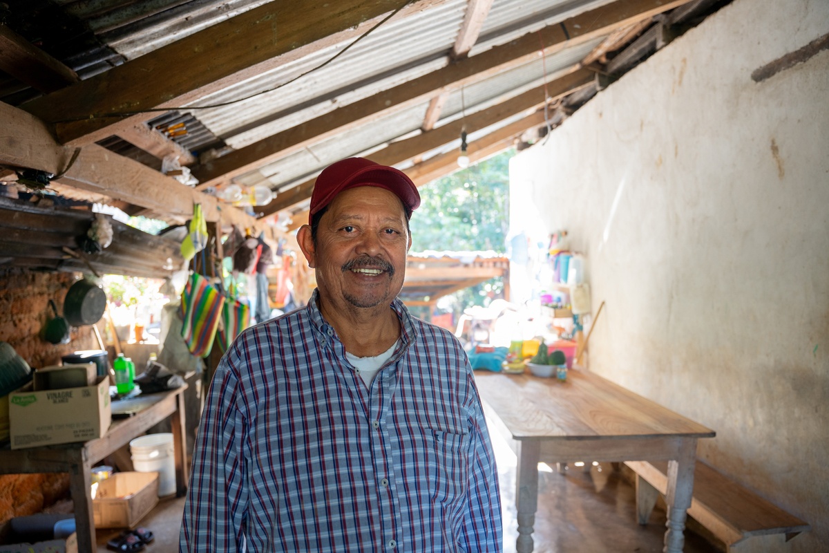 Carlos Vázquez stands in his home wearing a blue plaid shirt and a red baseball cap.
