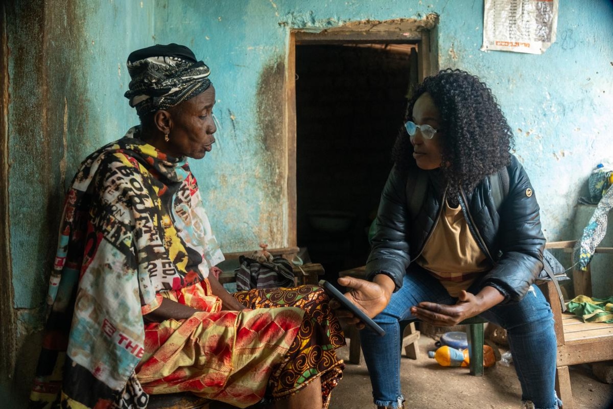Ramatu Jalloh, community health worker supervisor in Sierra Leone, sits with a patient in her home. Jalloh, who is wearing a leather jacket and jeans, is holding a smartphone, which she is using to track the home visit. Photo by Caitlin Kleiboer / PIH.