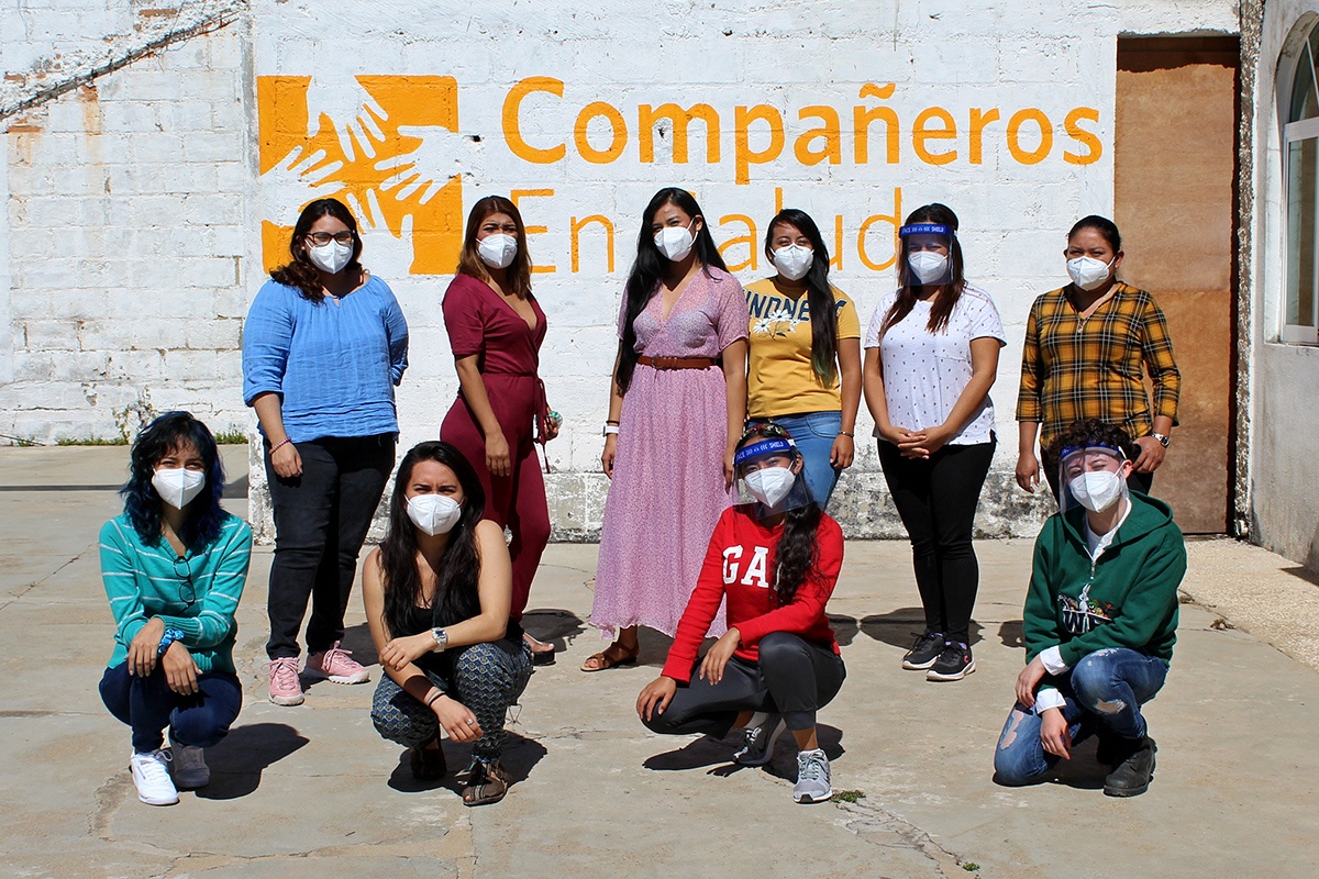 Ten women wearing face masks stand outside of a wall that says Compañeros En Salud, as Partners In Health is known in Mexico. Photo by Francisco Terán / Partners In Health.