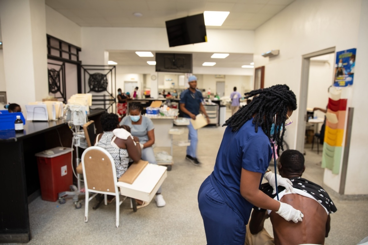A person in scrubs holds a stethoscope to someone's back with people in the background