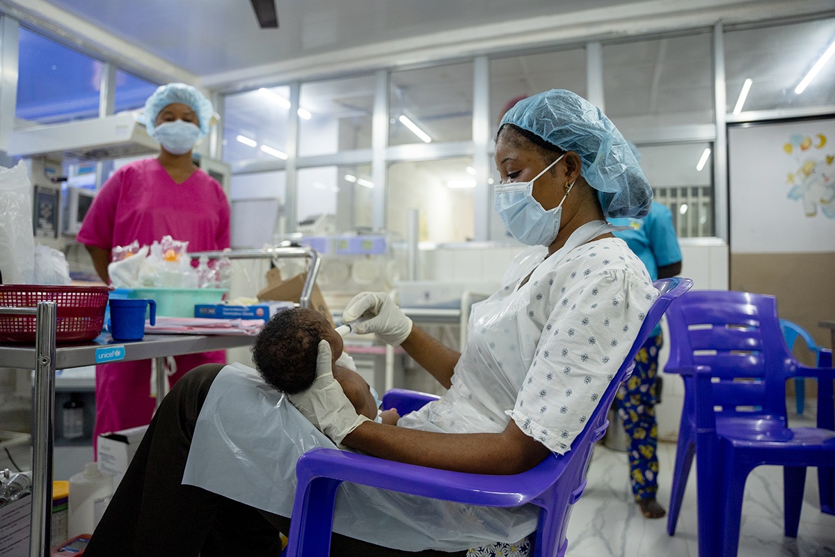 A nurse wearing a blue hair net and mask and a white plastic apron and gloves feeding a baby in the special care baby unit in Sierra Leone with a syringe
