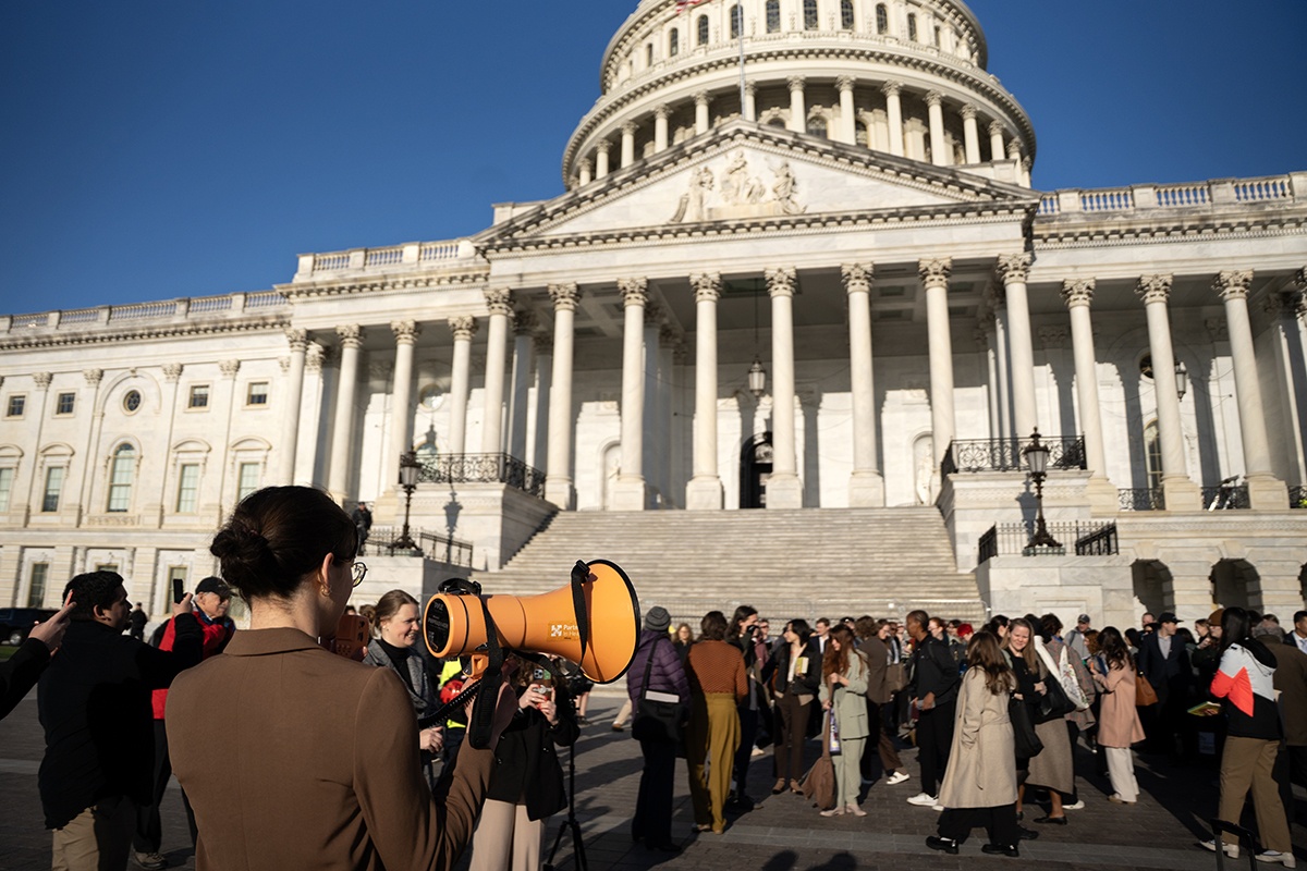 A large group of people gathered at the steps to the capitol being guided by a person holding an orange megaphone