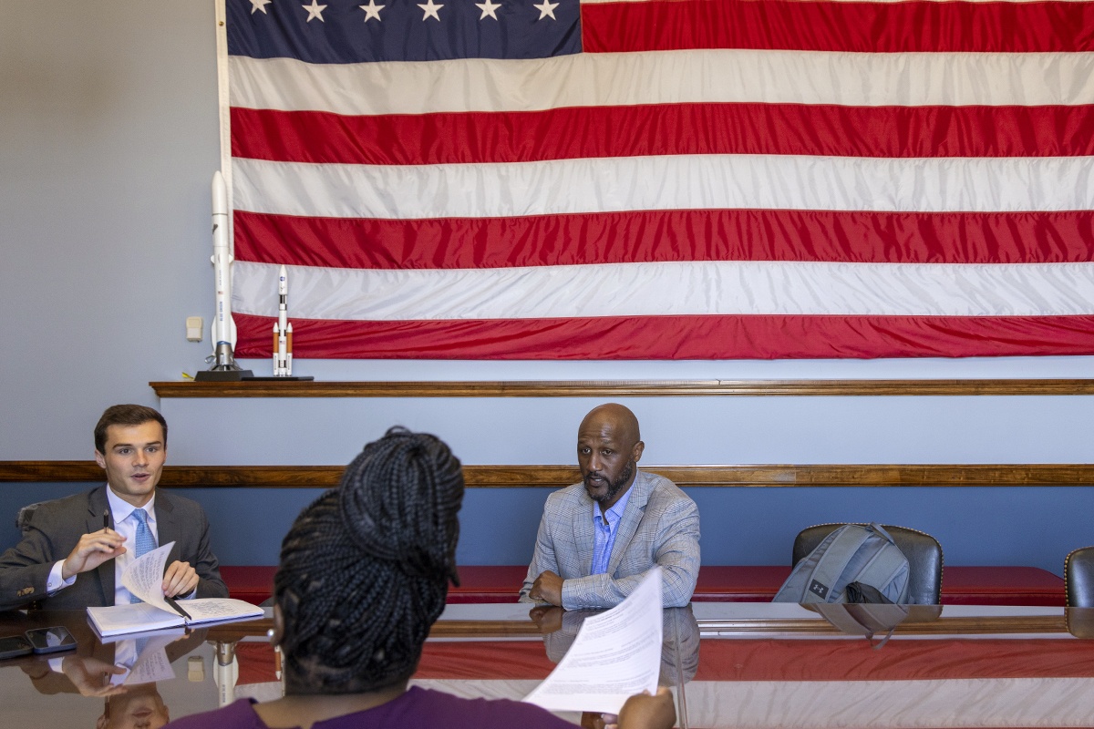 Three people talk with one another while sitting at tables underneath an American flag.