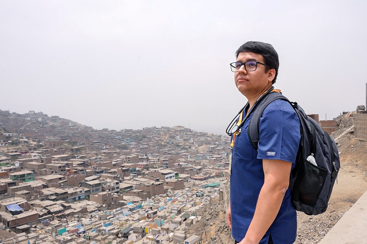 Dr. Santiago Palomino, wearing navy scrubs, a black backpack, black rimmed glasses, and a stethoscope around his neck, looks over a town from above
