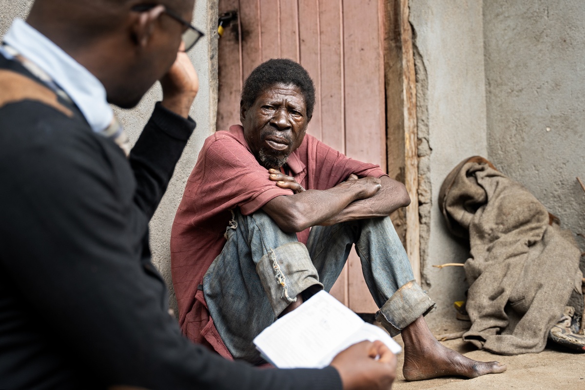 Saulos Metio, a man in his 60s, sits on the ground in a red shirt and blue jeans. He looks at another man, Charles Marshal, who is in the foreground of the photo.