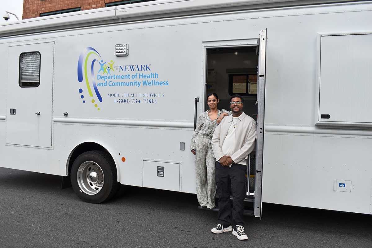 A large, white RV takes up the majority of the screen. In the RV's doorway stands a woman and a man. The woman has her arm propped on the man's shoulder.