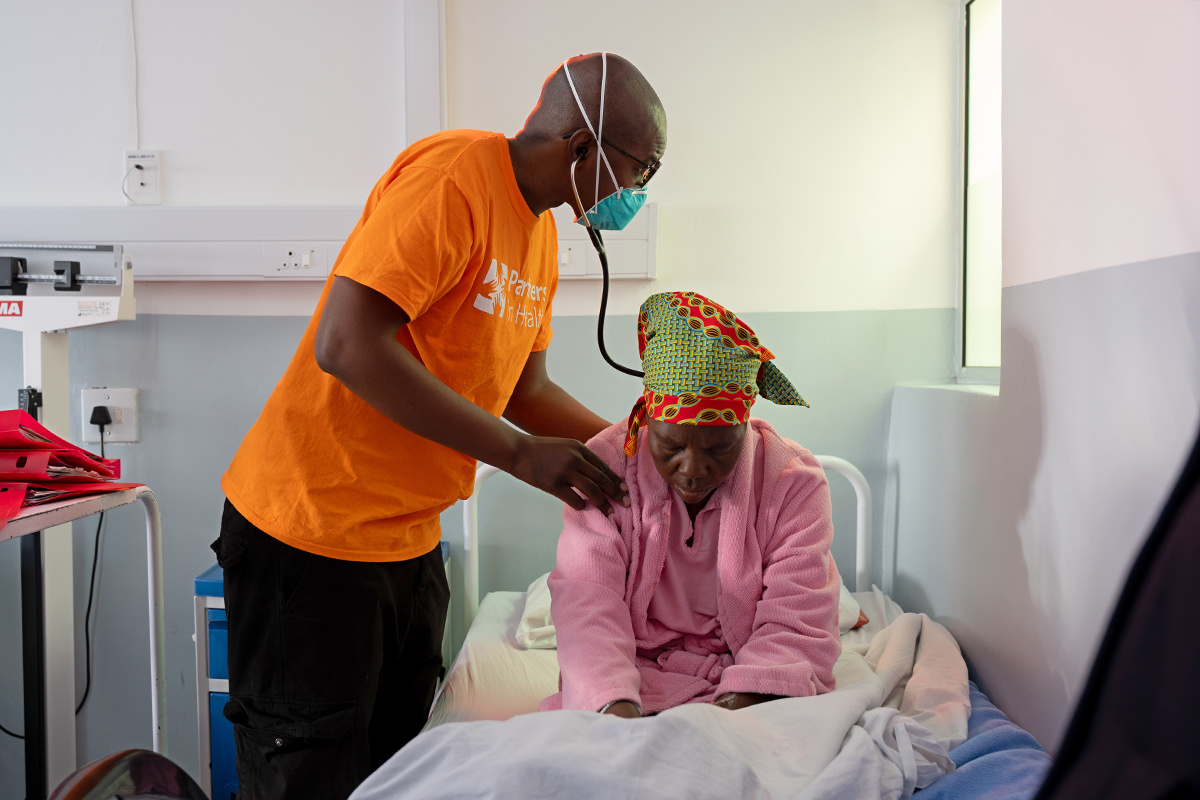 A person in an orange PIH shirt with a mask on holds a stethoscope to the back of a woman wearing pink sitting hunched over in a hospital bed.