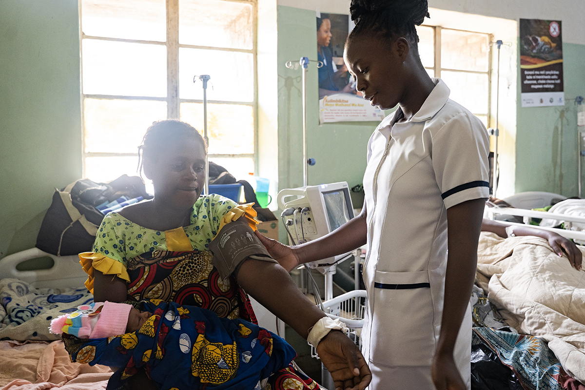 a nurse stands beside a partient sitting with a baby in her lap with a blood pressure cuff around her upper arm