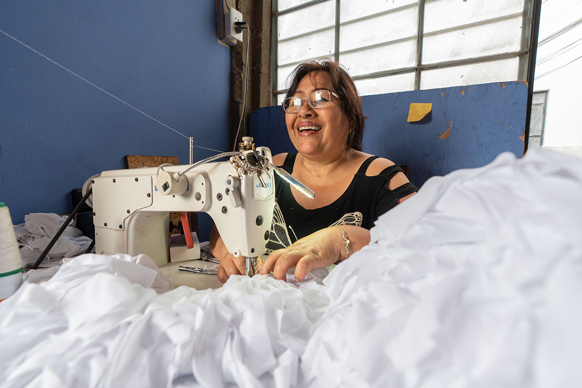María smiles while sitting at a sewing maching working with billowy white fabric