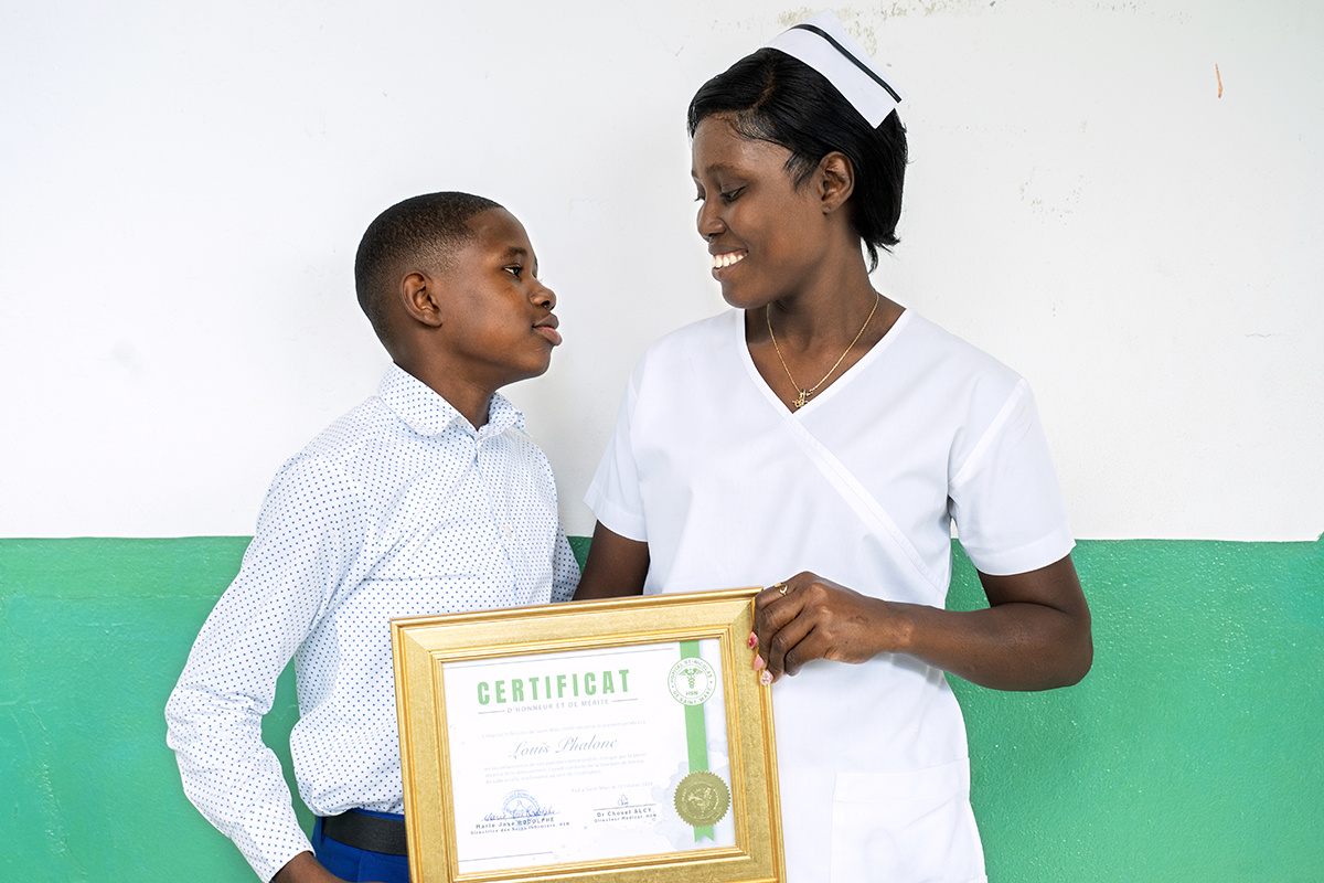 A woman in a nurse uniform smiles at her son while holding her nursing diploma.