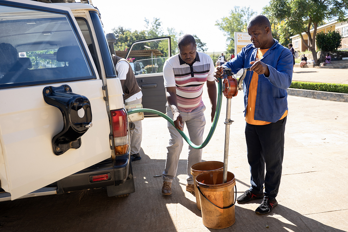 Two men stand to the right of a white van, filling it with fuel out of yellow buckets.