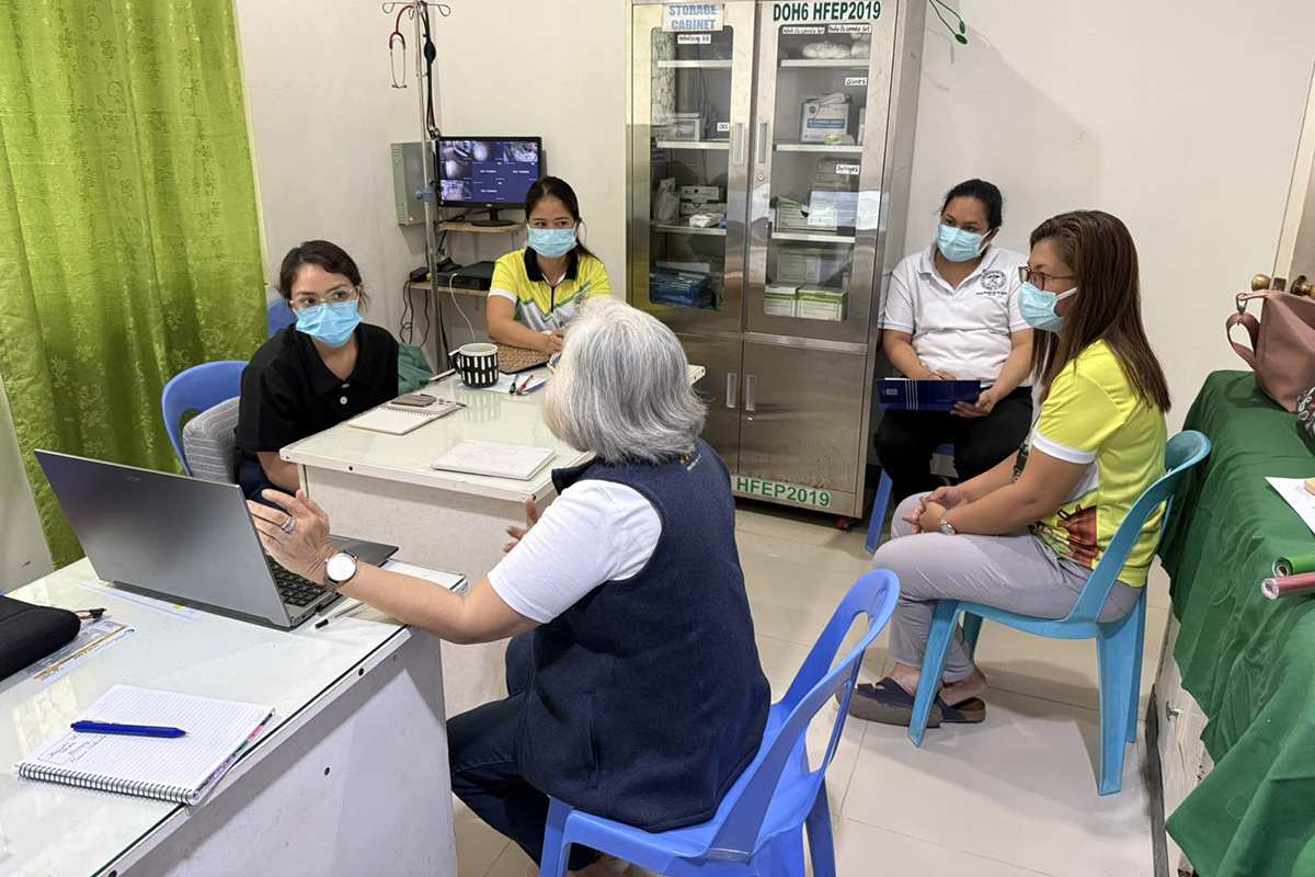 A group of health care workers sit around a table wearing masks. A person with a navy vest gestures toward a computer screen.