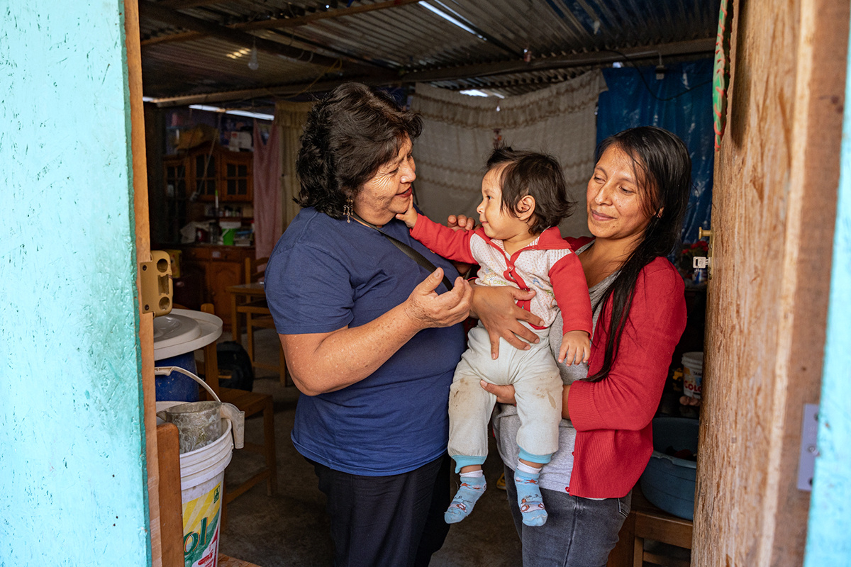 a woman in a red cardigan to the right holds a child in a white and red onesie who is reaching out to touch the cheek of a woman in a navy blue polo