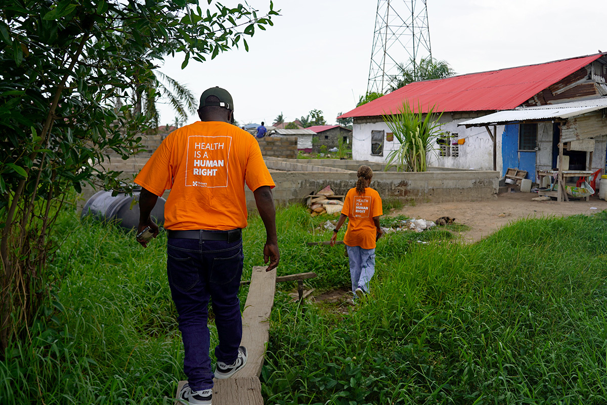 Two people walk along a path through grass to a home up ahead while wearing bright orange shirts that say "health is a human right"