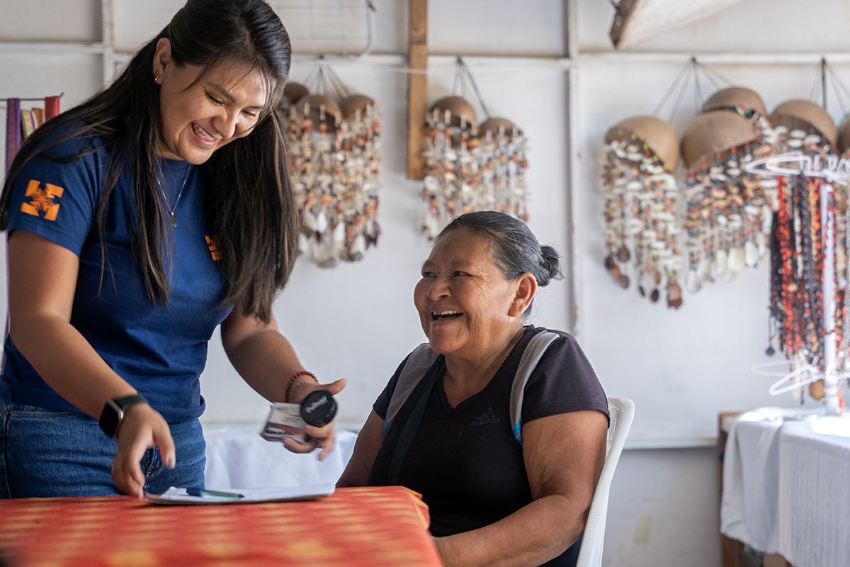 an older woman sits in a chair with a backpack on laughing while a younger woman in a navy blue shirt collects her forms, smiling with her