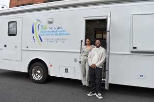 A large, white RV takes up the majority of the screen. In the RV's doorway stands a woman and a man. The woman has her arm propped on the man's shoulder.