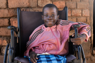 A young girl in a pink jacket smiles as she sits in a wheelchair. 