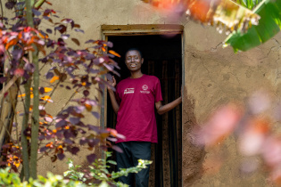 A young man stands in a doorway in a red shirt and smiles.