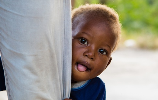 Lovenyou, a malnutrition patient, peeks out from his mother’s legs outside their home in Haiti. 