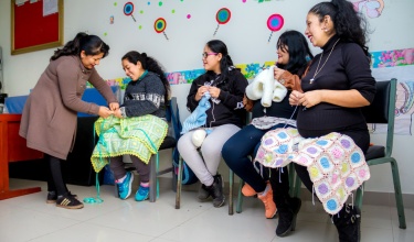 In Peru, mothers sit in a semicircle with knitted blankets and clothes on their laps as an instructor helps them.