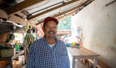 Carlos Vázquez stands in his home wearing a blue plaid shirt and a red baseball cap.