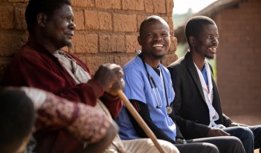 A PIH health worker in scrubs and with a stethoscope sits beside community members in Neno, Malawi.