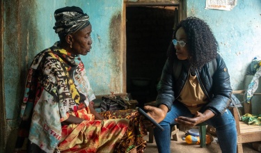 Ramatu Jalloh, community health worker supervisor in Sierra Leone, sits with a patient in her home. Jalloh, who is wearing a leather jacket and jeans, is holding a smartphone, which she is using to track the home visit. Photo by Caitlin Kleiboer / PIH.