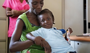 A young boy in a blue collared shirt smirking at the camera while being held by his mother who is smiling down at him