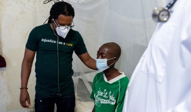 Dr. Maxo Luma, in a face mask and t-shirt that says "injustice has a cure," stands with a patient in a green shirt, also wearing a face mask.