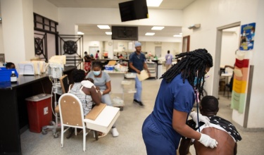 A person in scrubs holds a stethoscope to someone's back with people in the background