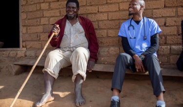 A man in a red jacket sits while holding a walking stick. Beside him is a man in scrubs with a stethoscope around his neck.