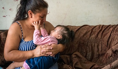 A mother in a striped dress holds her daughter in her arms as the child grabs her mother's cheeks