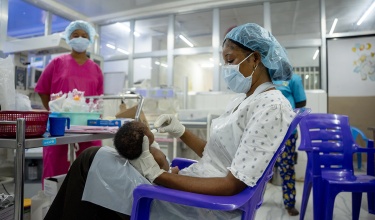 A nurse wearing a blue hair net and mask and a white plastic apron and gloves feeding a baby in the special care baby unit in Sierra Leone with a syringe